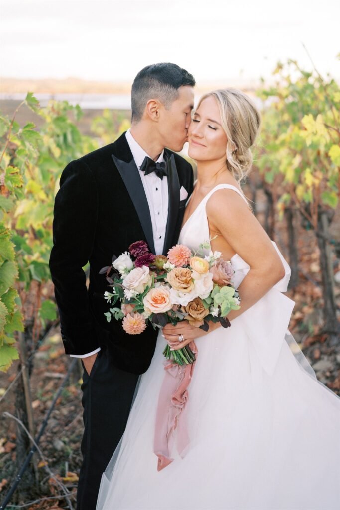 A groom in a black tuxedo kisses a smiling bride holding a bouquet. They stand together in a vineyard, surrounded by rows of green grapevines.
