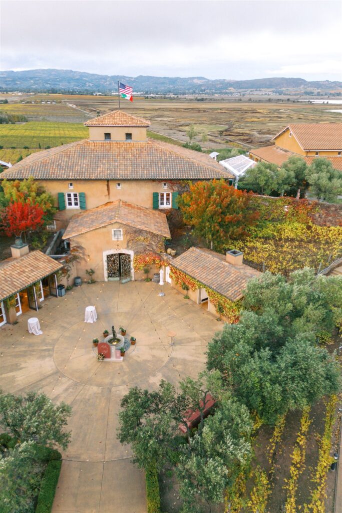 Aerial view of a large, rustic building with a circular courtyard and American flag on the roof, surrounded by vineyards and hills in the background.