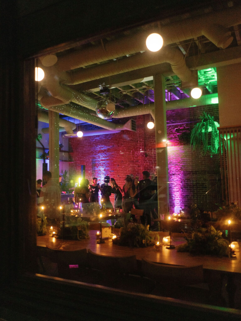 View through a window of a dimly lit room with people gathered, colorful lighting on the walls and ceiling, a disco ball, and tables decorated with plants and candles.