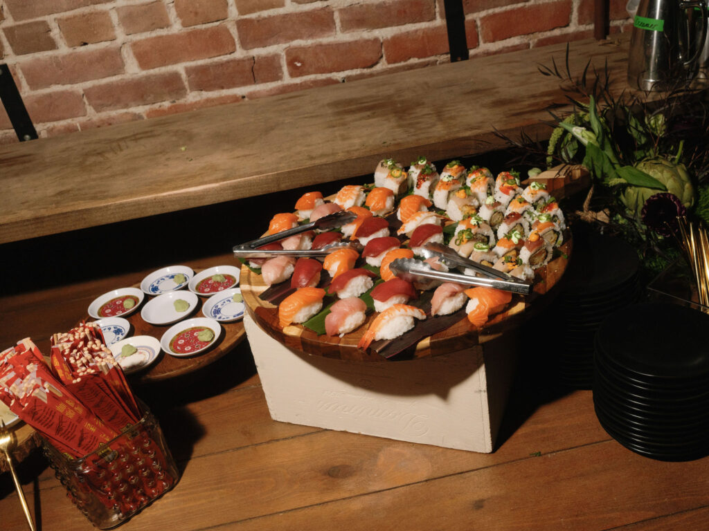 A wooden platter with assorted sushi, including nigiri and rolls, is on a table alongside chopsticks and small soy sauce packets.