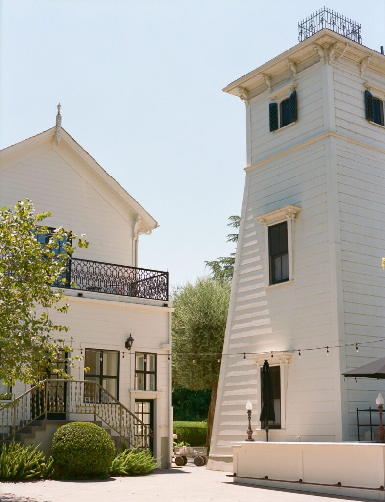 Two white wooden buildings with windows and a balcony, separated by a paved area. The left building has stairs, and string lights hang in the foreground. Trees and a clear sky are visible.