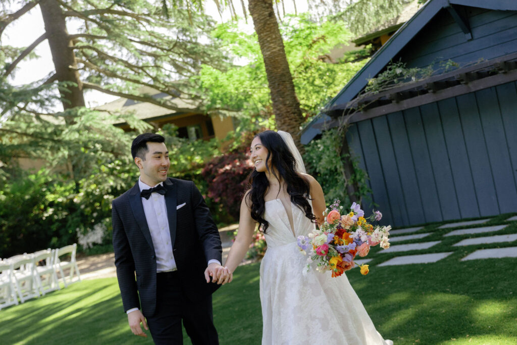 A bride in a white dress holding a colorful bouquet walks hand in hand with a groom in a black suit on a lawn, both smiling at each other.