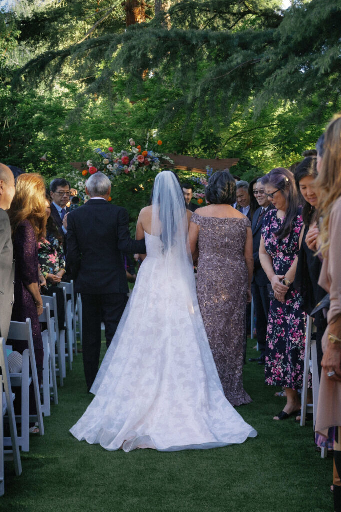 A bride in a white dress walks down an outdoor aisle with two people, surrounded by seated guests under trees and floral decorations.
