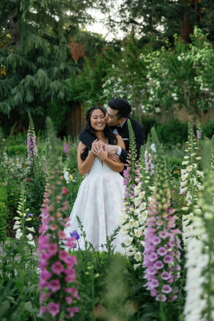 A couple in wedding attire embraces and smiles while standing in a garden surrounded by tall flowers and greenery.