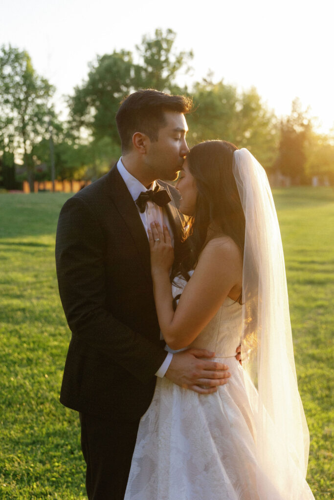 A groom in a dark suit kisses the forehead of a bride in a white dress and veil as they embrace outdoors at sunset.