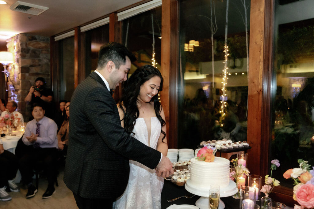 A couple in formal attire cuts a white tiered cake together at a wedding reception, surrounded by candles and cupcakes, with guests seated in the background.
