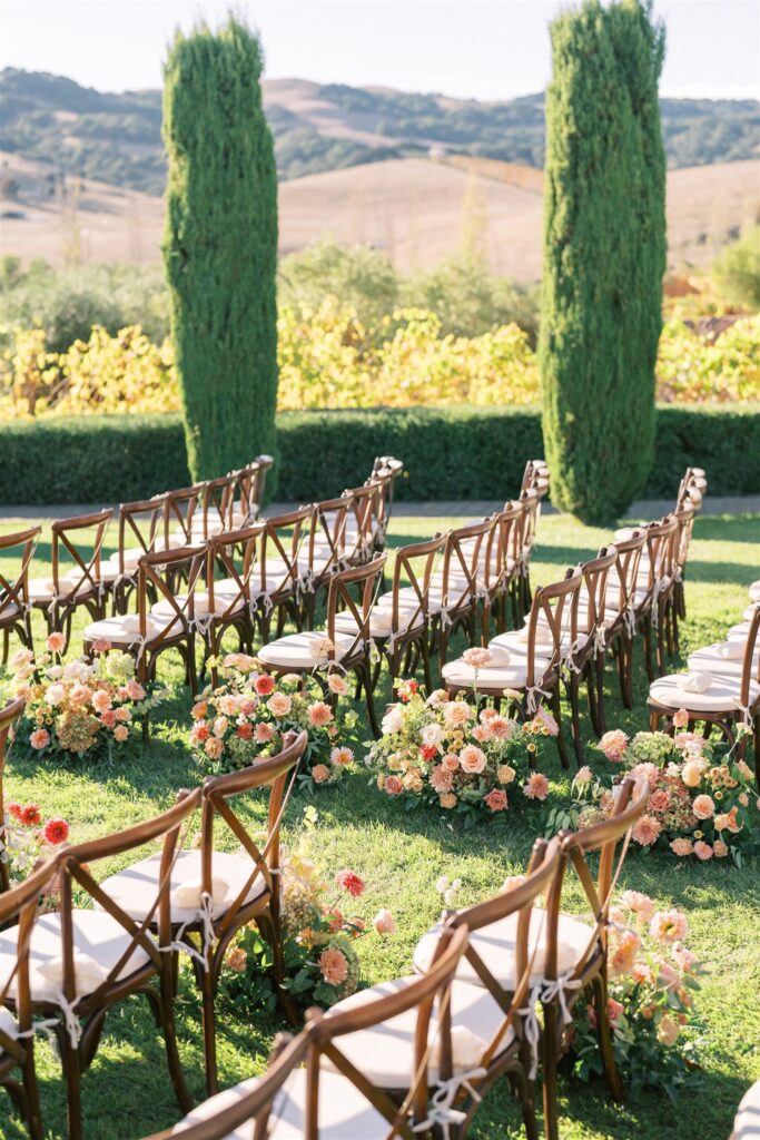 An outdoor wedding setup with rows of wooden chairs facing forward. The aisle is lined with flower arrangements, and tall trees and rolling hills are visible in the background.
