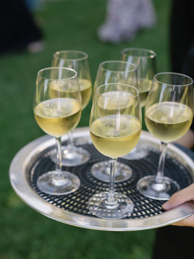 A tray carrying six glasses of white wine is held outdoors on a grassy area.