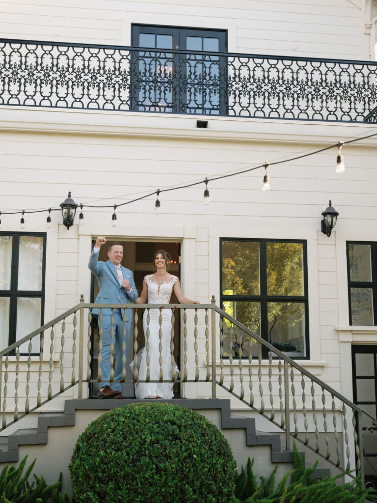 A couple stands on a staircase outside a white building with a black railing. The man gestures with his hand while the woman smiles, adorned in formal attire. Hanging lights and greenery decorate the scene.