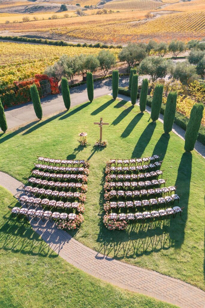 Outdoor wedding ceremony setup with rows of chairs facing a wooden cross and a small table on a green lawn, surrounded by tall trees and extensively cultivated fields in the background.