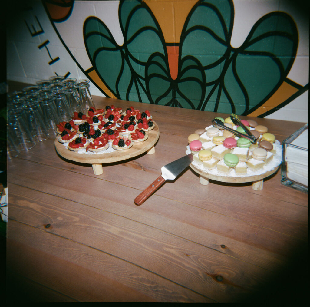A wooden table with two dessert platters: one with assorted berries and another with colorful macarons. A knife and stack of napkins are nearby. Glasses are lined up on the side.