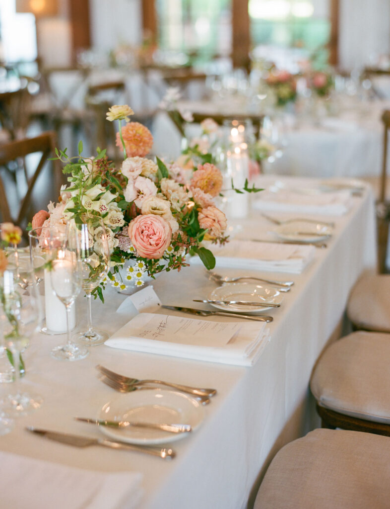 Elegant table setting with white tablecloths, floral centerpieces, glassware, and silverware. Plates are neatly arranged with napkins and menus.