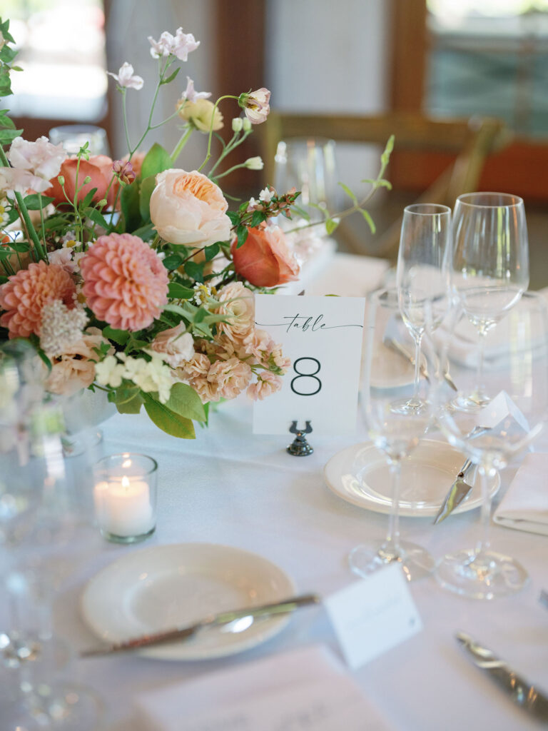 Table setting featuring pink and peach flowers, candles, and a table number card labeled "Table 8" on a white tablecloth.
