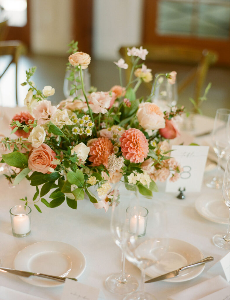 A floral centerpiece with pink and peach flowers on a white tablecloth. Surrounding the arrangement are empty wine glasses, small candles, and a table number card.