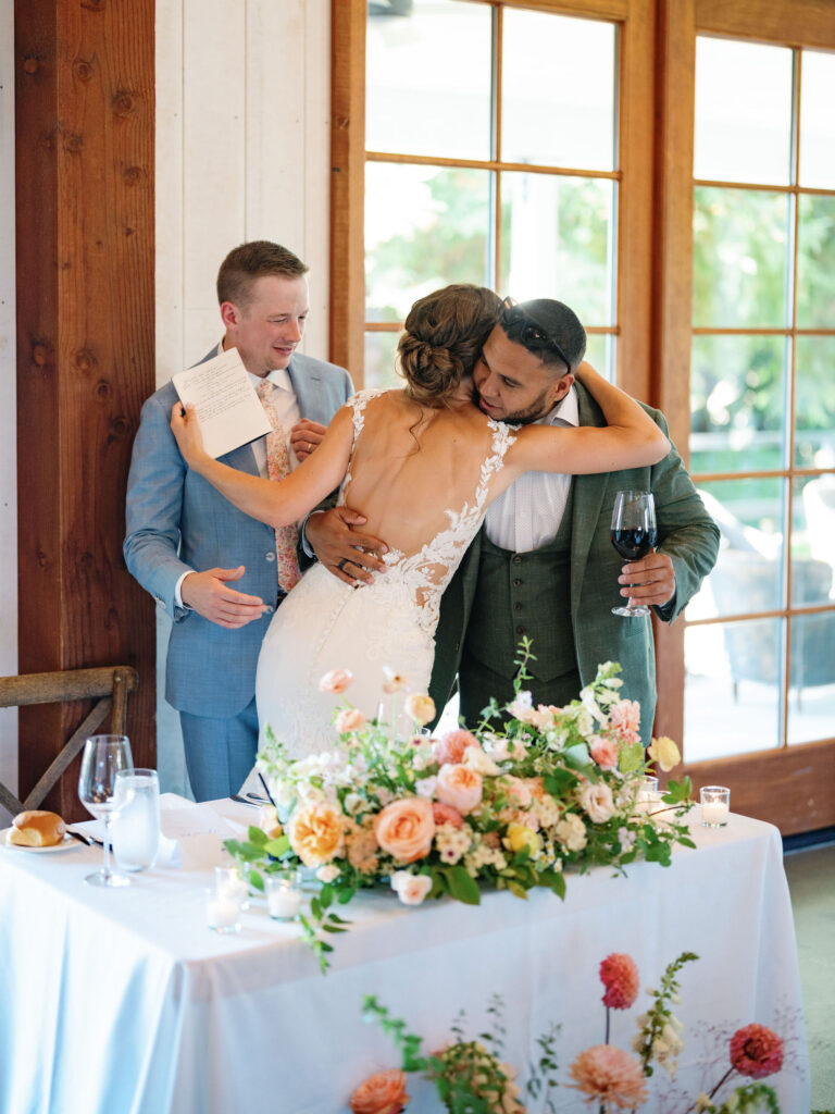 Three people at a wedding reception. The bride hugs a man in a dark suit holding a wine glass, while another man in a light blue suit smiles, holding a paper. A table with flowers is in front of them.