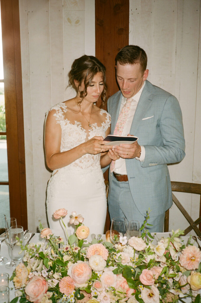 Bride and groom in formal attire looking at a tablet, standing behind a table with floral arrangements.