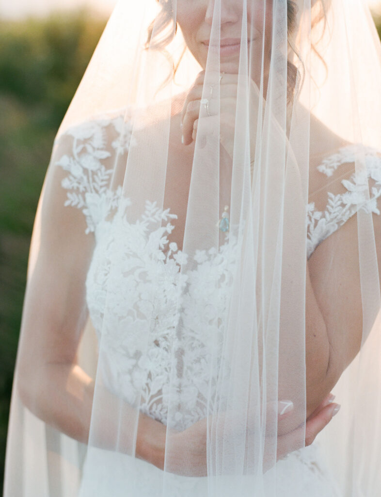 Bride in a lace wedding dress partially obscured by a sheer veil, standing outdoors with her hand gently touching her chin.