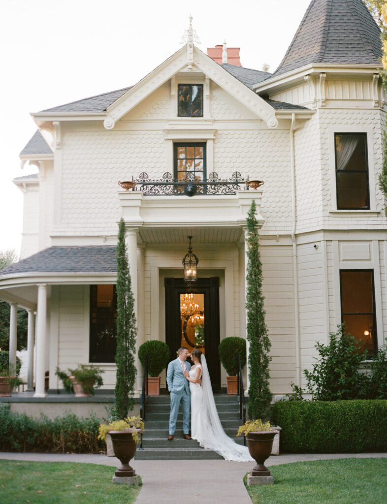 A couple in wedding attire stands on the steps of a large Victorian-style house with a pointed roof and decorative balcony. Tall plants and a manicured garden surround the entrance.