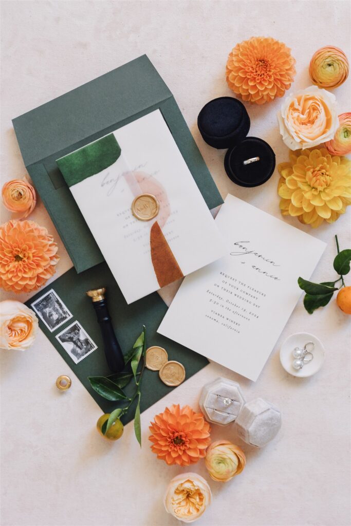 A wedding invitation suite with green and white cards, wax seal, stamps, rings in black and gray boxes, surrounded by orange and yellow flowers and green leaves.