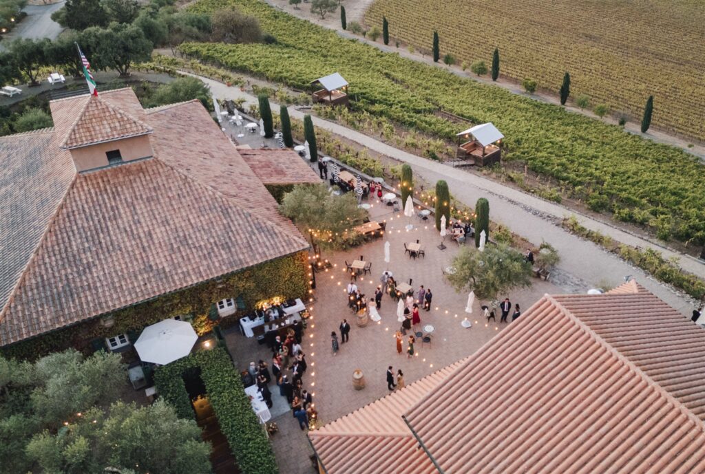 Aerial view of a courtyard event with guests mingling between rustic buildings surrounded by vineyards and trees.