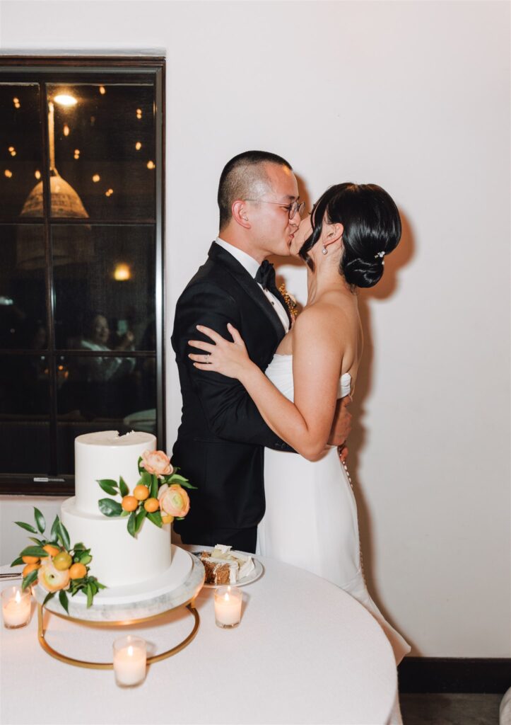 Couple in formal attire kisses beside a white wedding cake adorned with orange flowers and greenery.