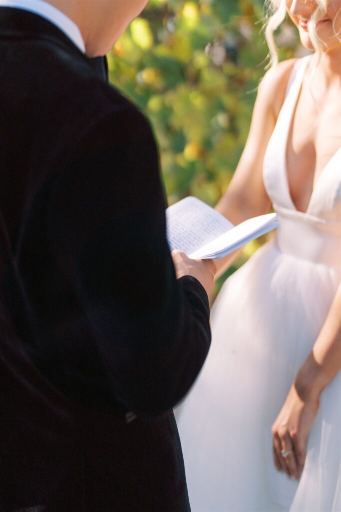 A person in a dark suit reads from a notebook while a person in a white dress listens, set against a background of green foliage.