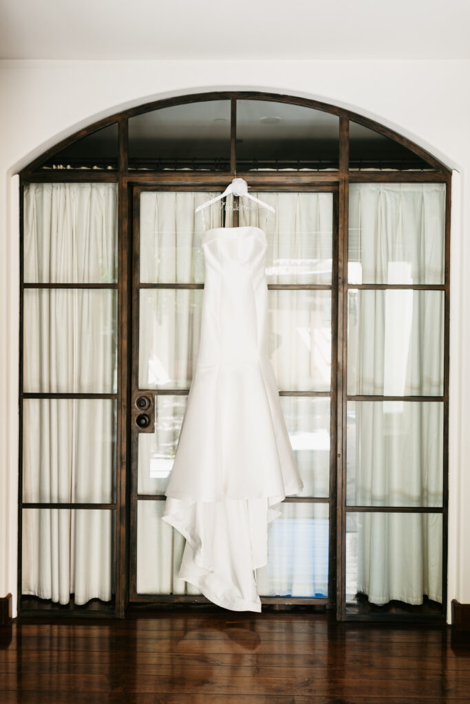 A white strapless wedding dress hangs on a wooden hanger in front of a glass-paneled door with white curtains.