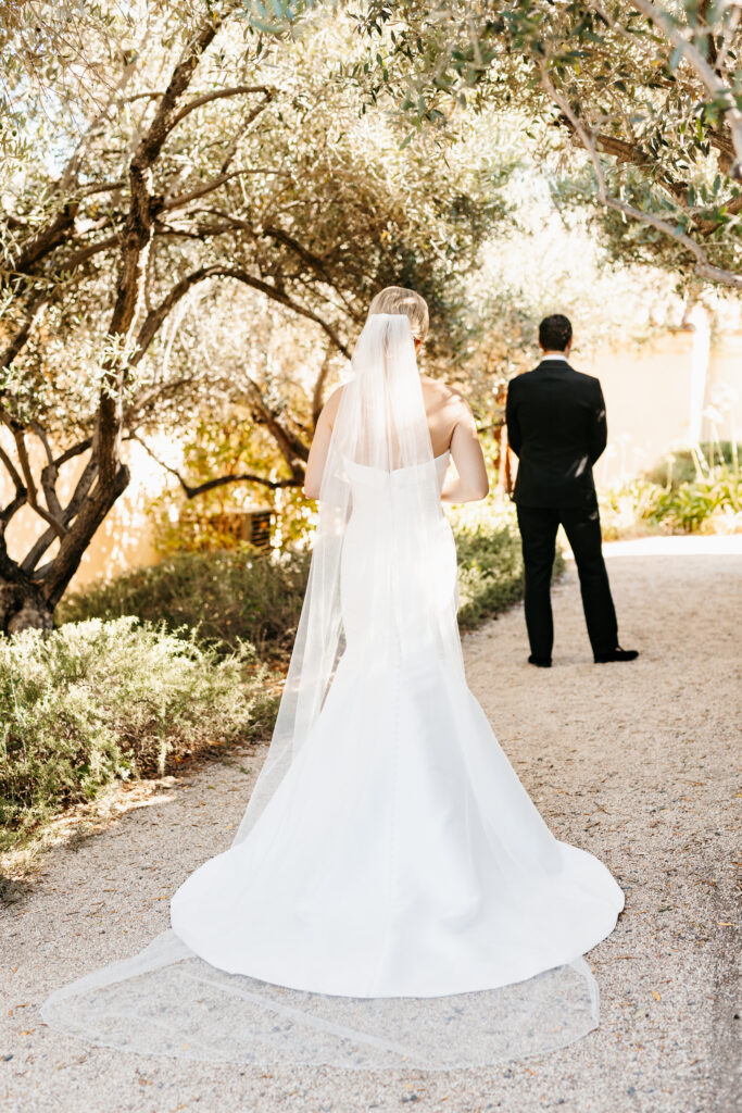A bride in a white dress and veil walks toward a man in a suit, standing on a sunlit garden path surrounded by trees.