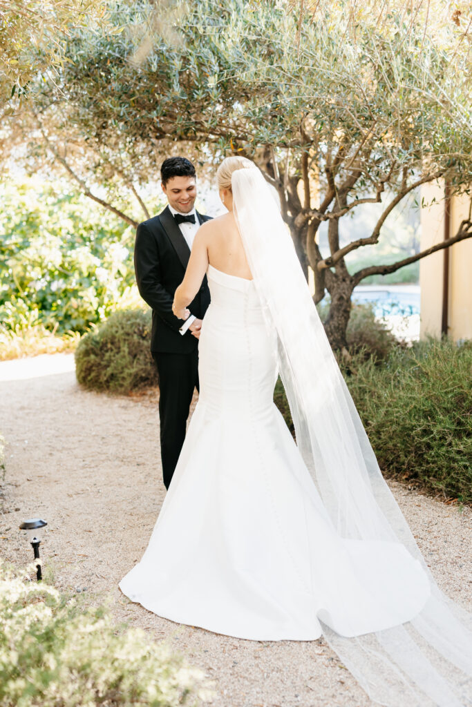 A bride in a white strapless gown and long veil holds hands with a groom in a black tuxedo outdoors under leafy trees.