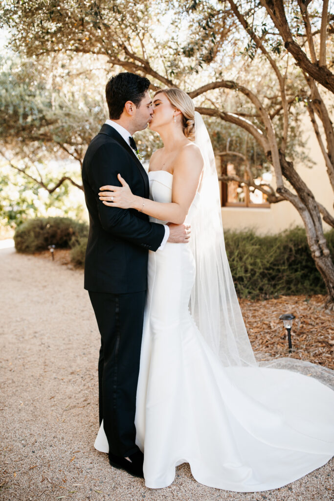 Bride and groom in formal attire share a kiss outdoors under trees on their wedding day.