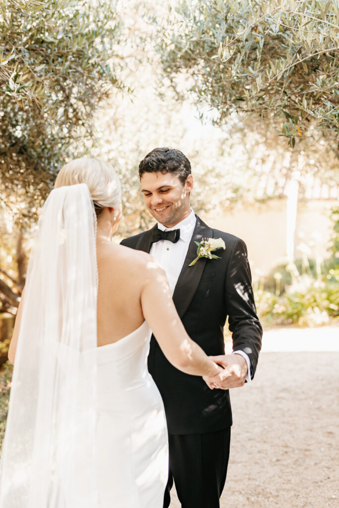 A bride in a white dress and veil holds hands with a groom in a black tuxedo outdoors, surrounded by greenery.