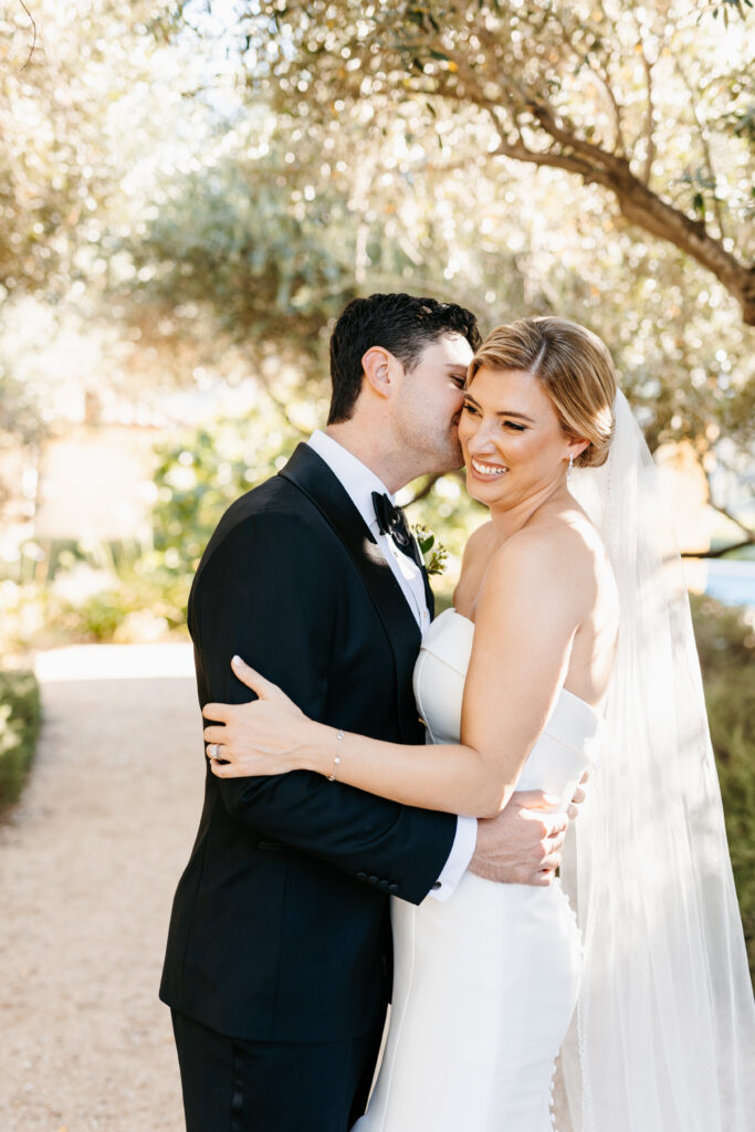 A bride in a strapless white dress and veil smiles as the groom in a black tuxedo embraces and kisses her on the cheek outdoors.