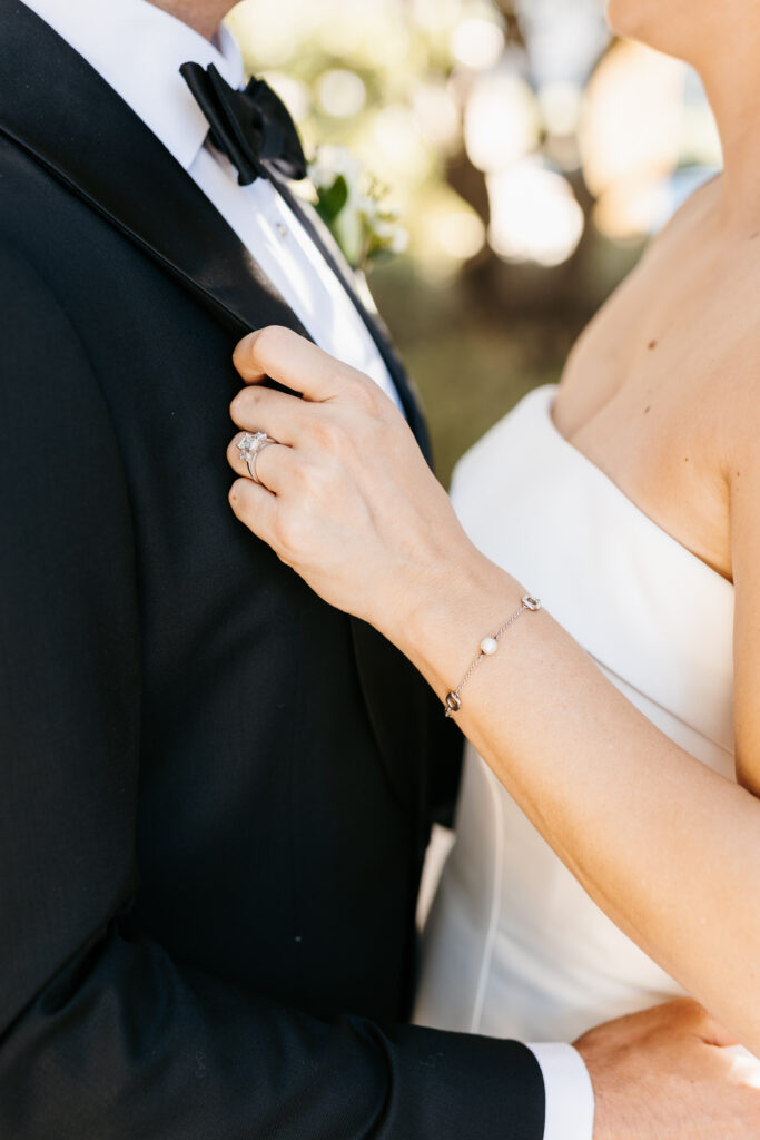 A bride in a strapless white dress holds the lapel of a groom's black tuxedo jacket, showing her wedding ring and bracelet.