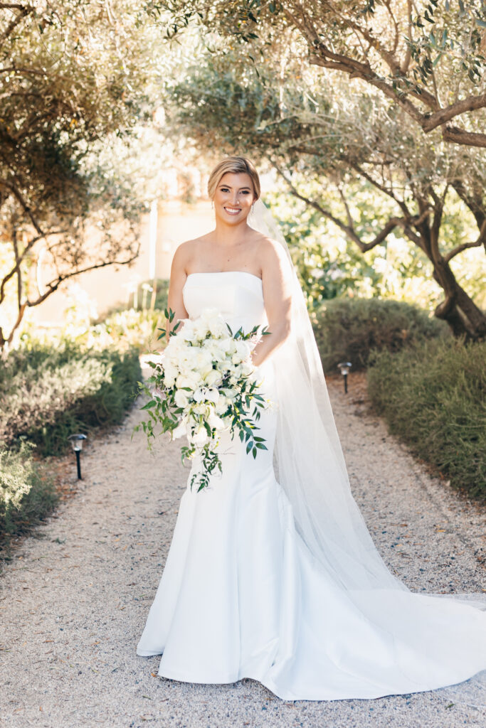 A bride in a strapless white wedding dress holds a large bouquet of white flowers while standing on a gravel path surrounded by greenery.