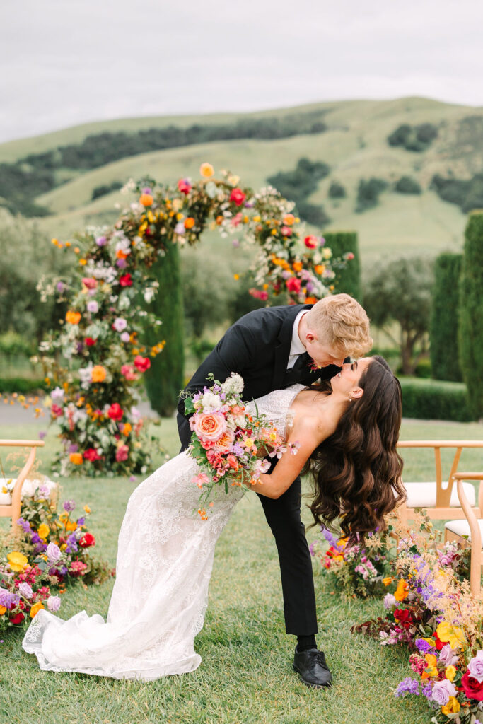 A groom kisses a bride while dipping her backward. They're outdoors, surrounded by colorful flowers, with rolling hills in the background.