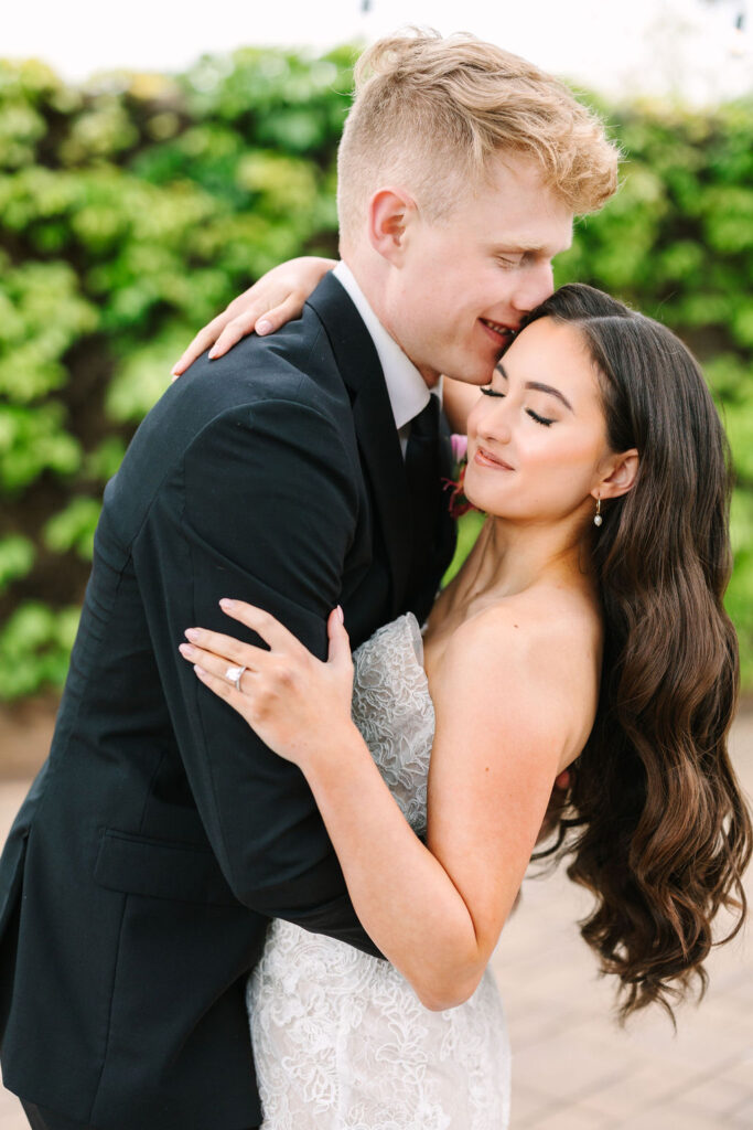 A couple embraces outdoors. The man in a black suit leans in, and the woman in a strapless white dress smiles with closed eyes. Green foliage is in the background.
