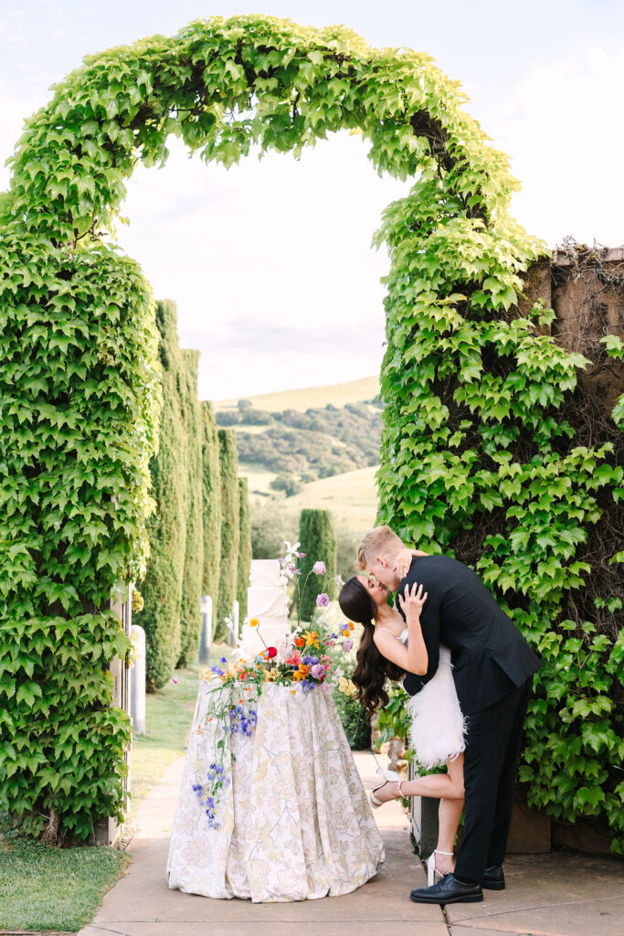 A couple shares a kiss under a tall, greenery-covered archway. A small table with a floral arrangement stands beside them, with hills visible in the background.