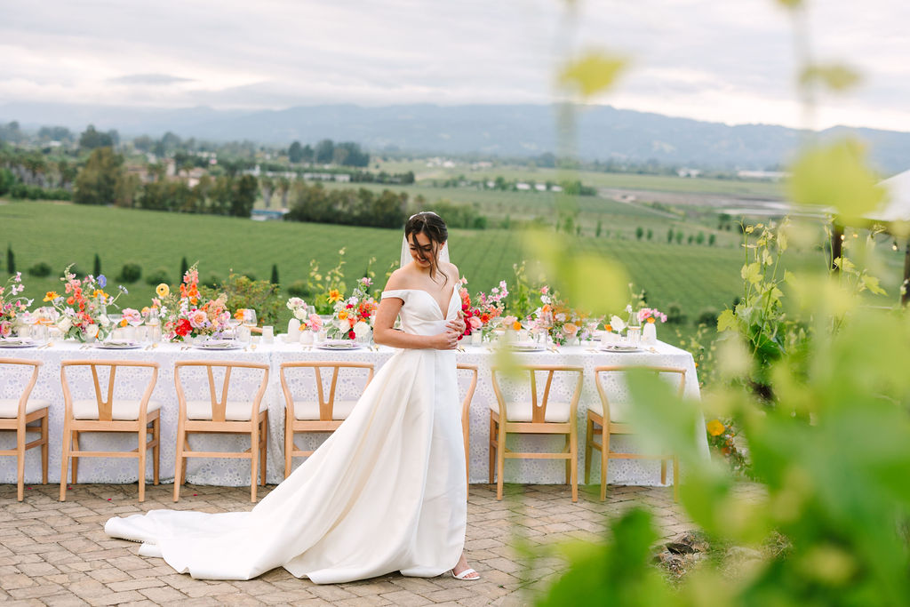 Bride in a white dress stands beside a long table adorned with colorful flowers, set against a backdrop of vineyards and hills under a cloudy sky.
