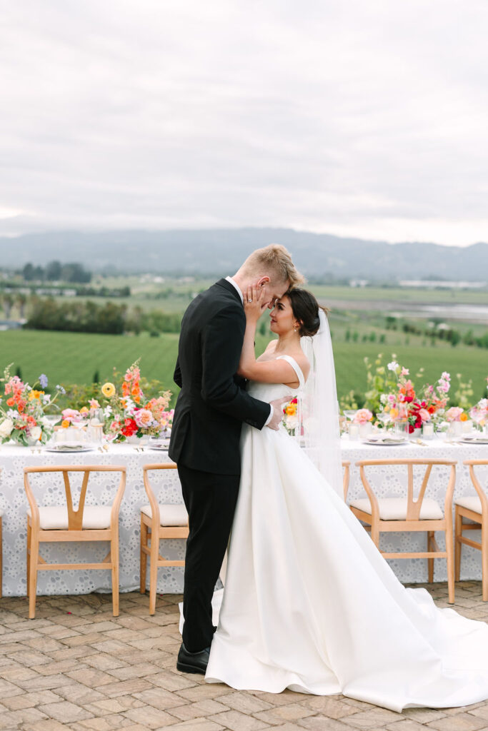 A bride and groom share a moment in front of a decorated table with colorful flowers, set outdoors against a scenic landscape with cloudy skies.