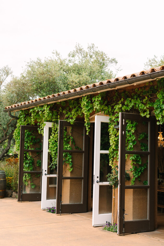 Large open doors surrounded by green ivy on a rustic building with a tile roof, set against a background of trees.