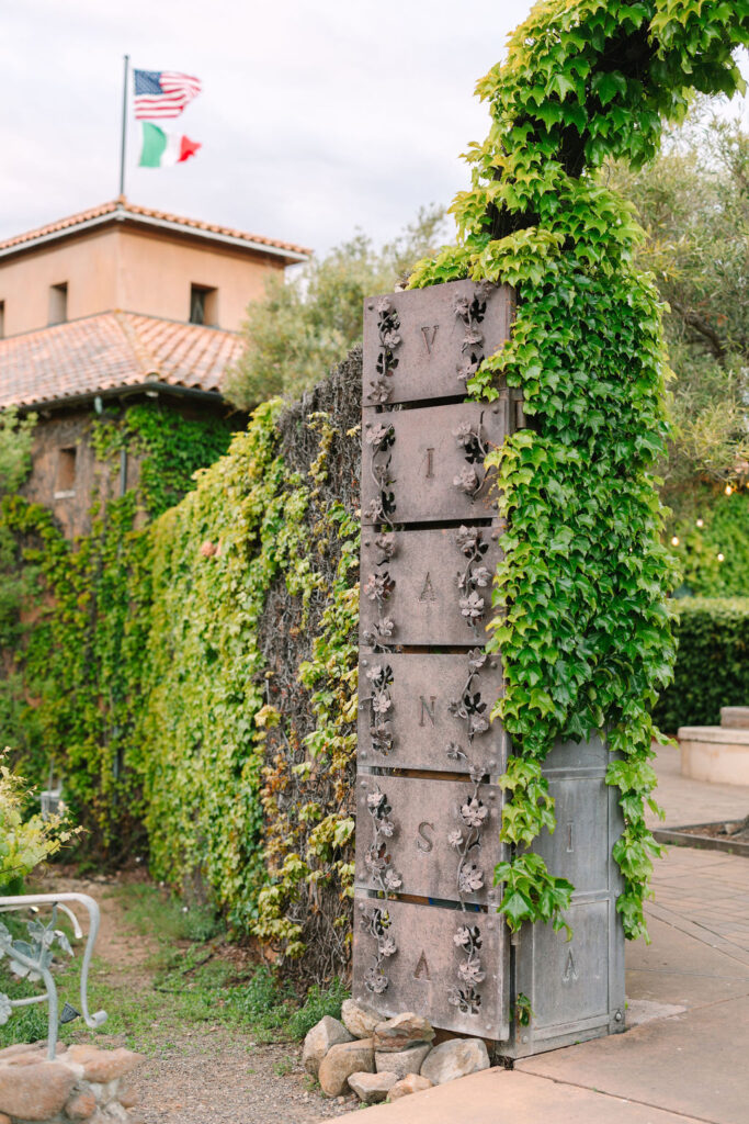 A vertical stone sign with the word "VIANSA" is covered in green ivy. Behind it is a building with U.S. and Italian flags on top.
