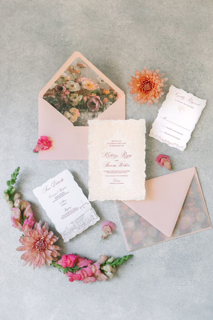 A wedding invitation set with floral designs, including envelopes and RSVP cards, arranged on a textured surface, accented with pink and orange flowers.