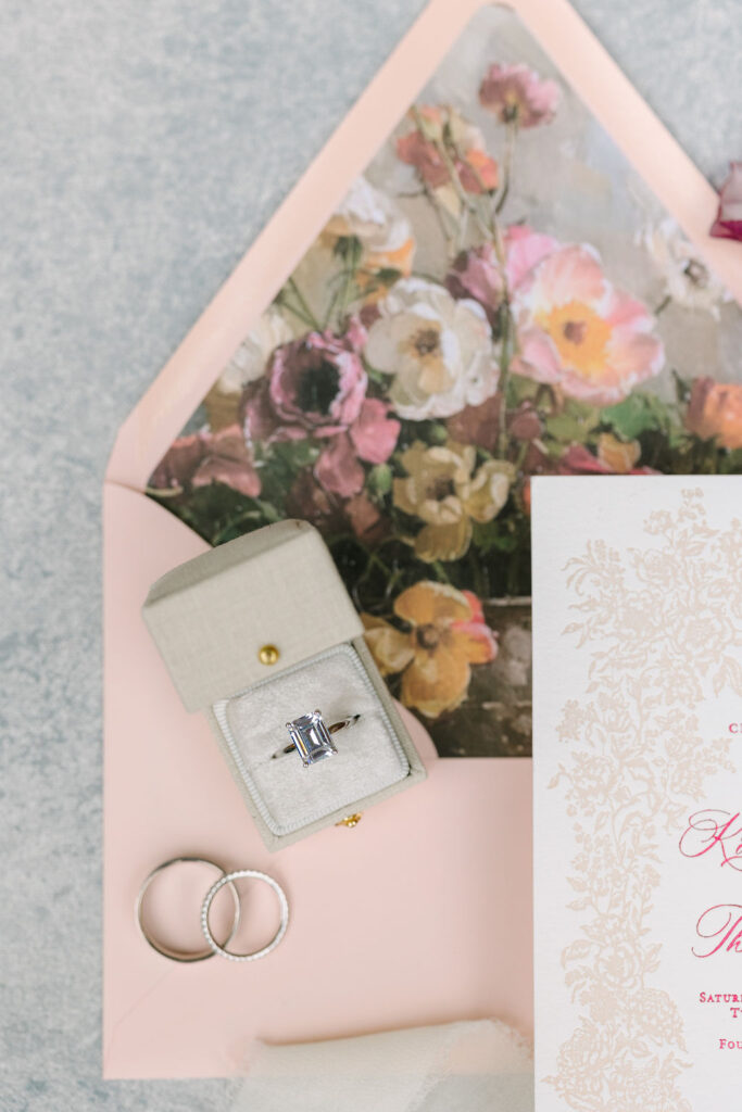 An engagement ring in a box, two wedding bands, a floral envelope, and a decorative invitation are arranged on a textured surface.