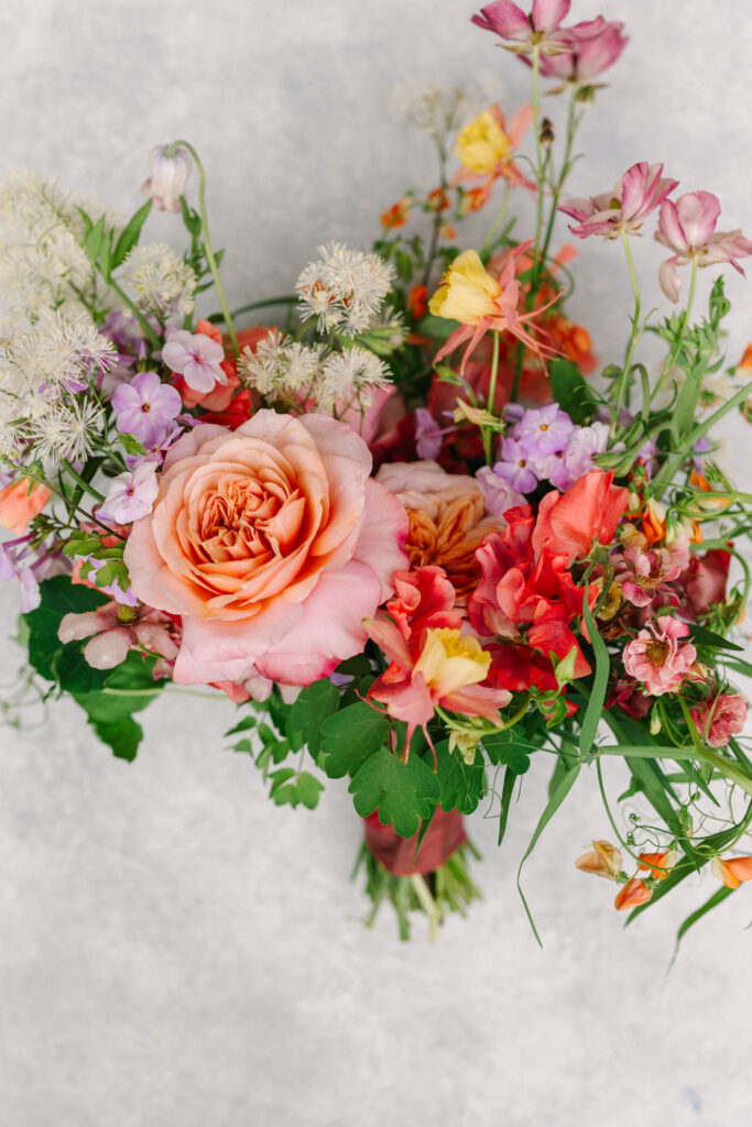 Bouquet of various colorful flowers, including a large peach rose, red and pink blooms, and green foliage, arranged against a light background.