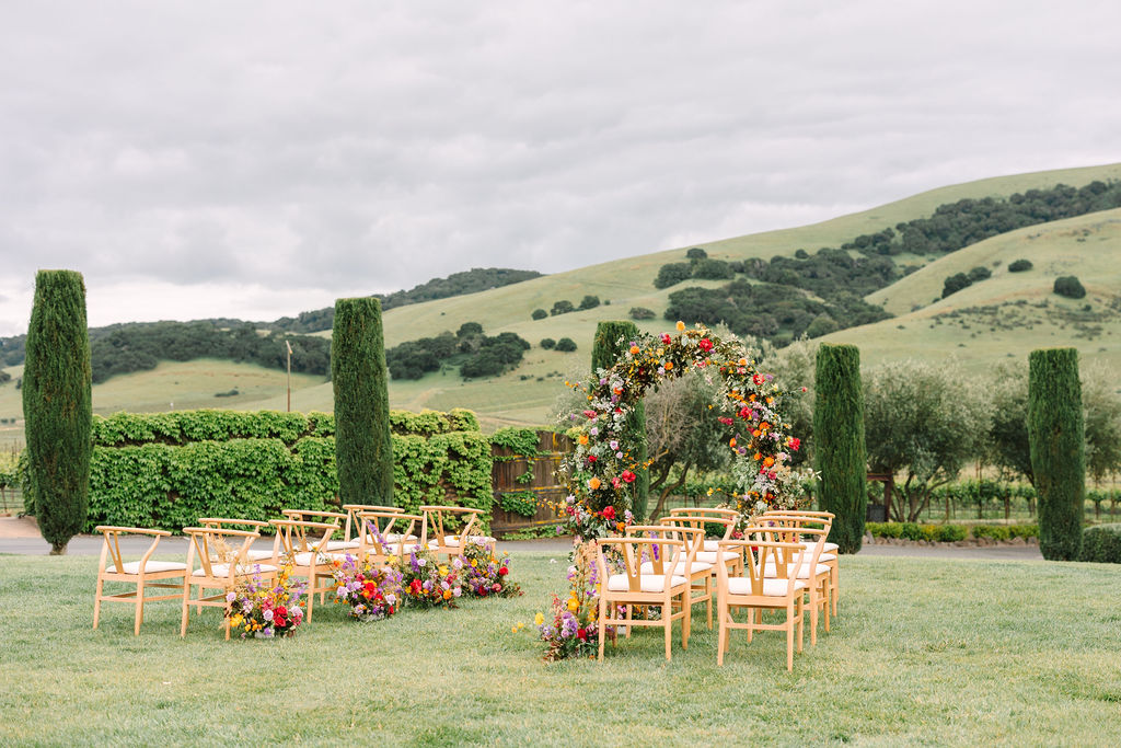 Outdoor wedding setup with wooden chairs facing a circular floral arch adorned with colorful flowers, set against a backdrop of rolling green hills and tall trees under a cloudy sky.