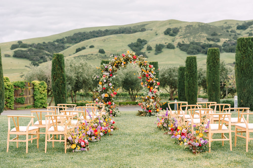 Outdoor wedding setup with wooden chairs facing a circular floral arch, surrounded by colorful flowers. Green hills and tall trees are in the background under a cloudy sky.