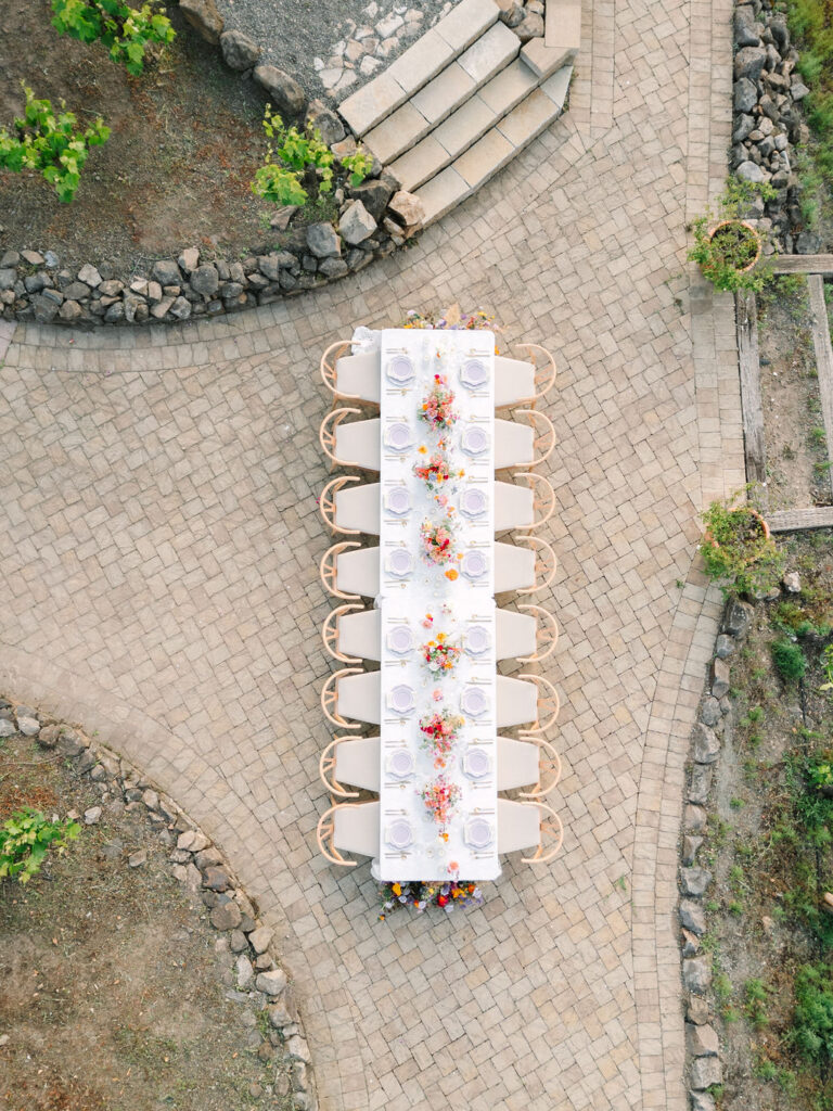Aerial view of an outdoor dining table set with white linens, plates, and floral centerpieces, placed on a stone courtyard with surrounding greenery.