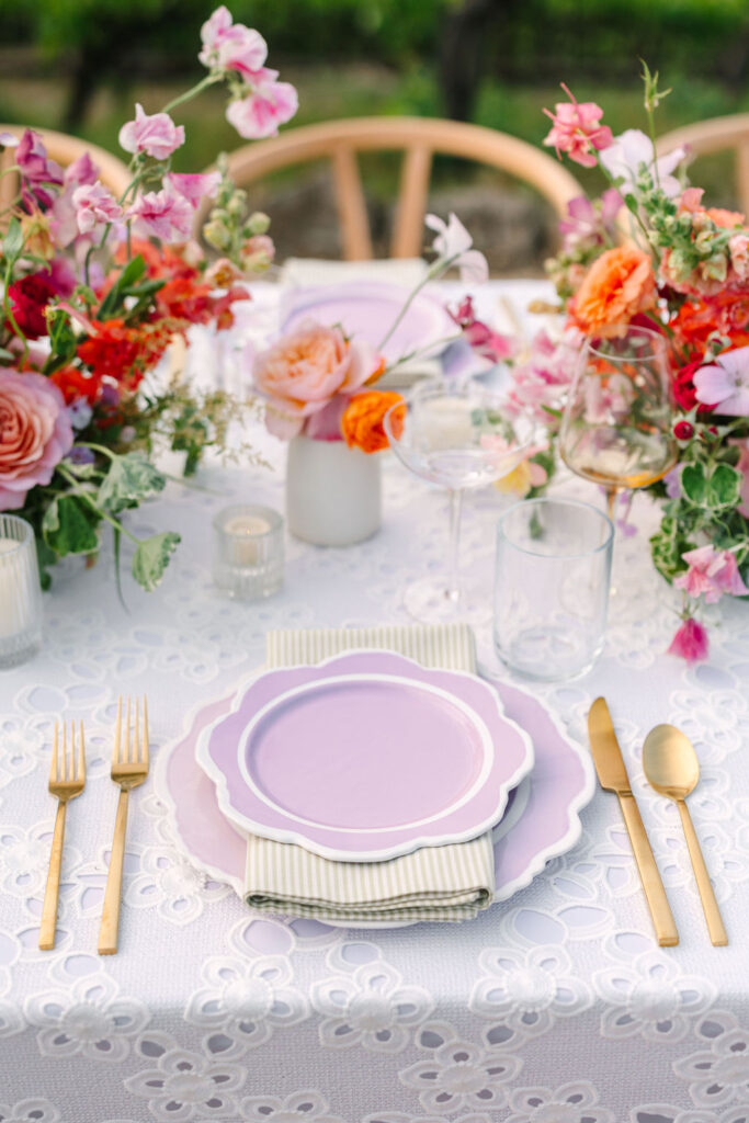 Elegant table setting with a lavender plate on a patterned napkin, gold cutlery, glassware, and colorful floral centerpieces on a white lace tablecloth.