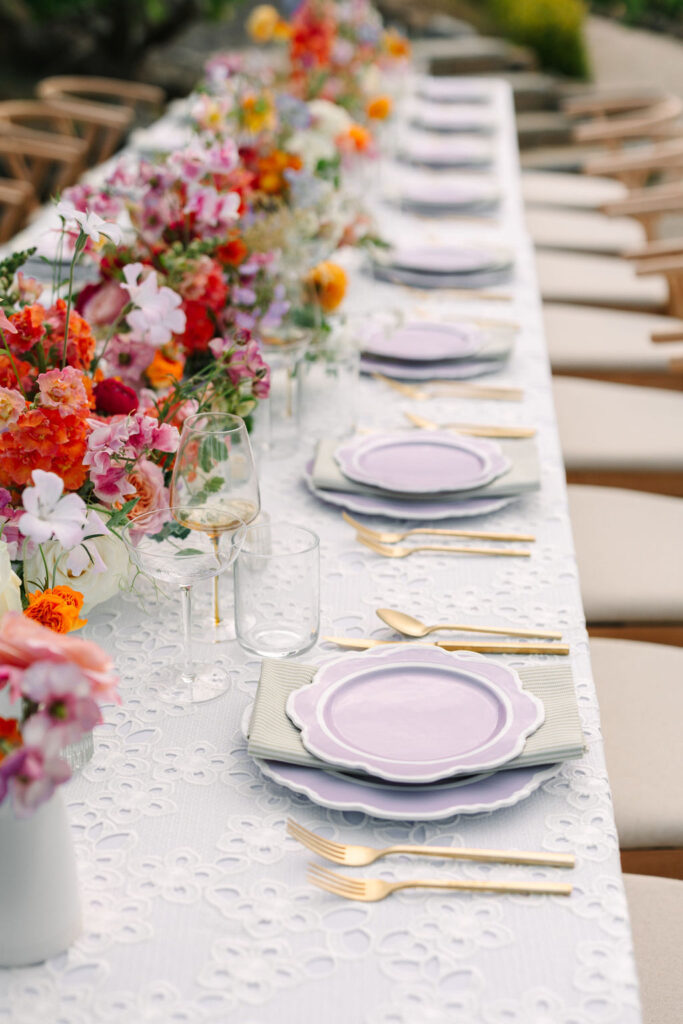 Elegant outdoor dining table set with purple plates, gold cutlery, and floral centerpieces with pink, orange, and red flowers. White lace tablecloth and wooden chairs are arranged neatly.