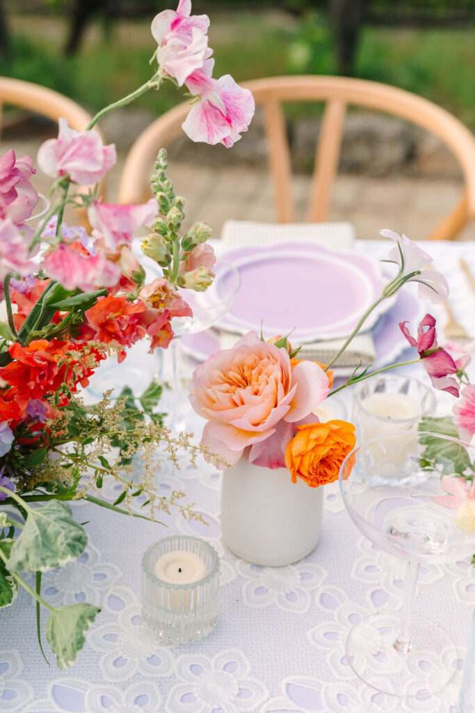 Floral table setting with colorful flowers in vases, a purple plate, and a lit candle on a lace tablecloth.